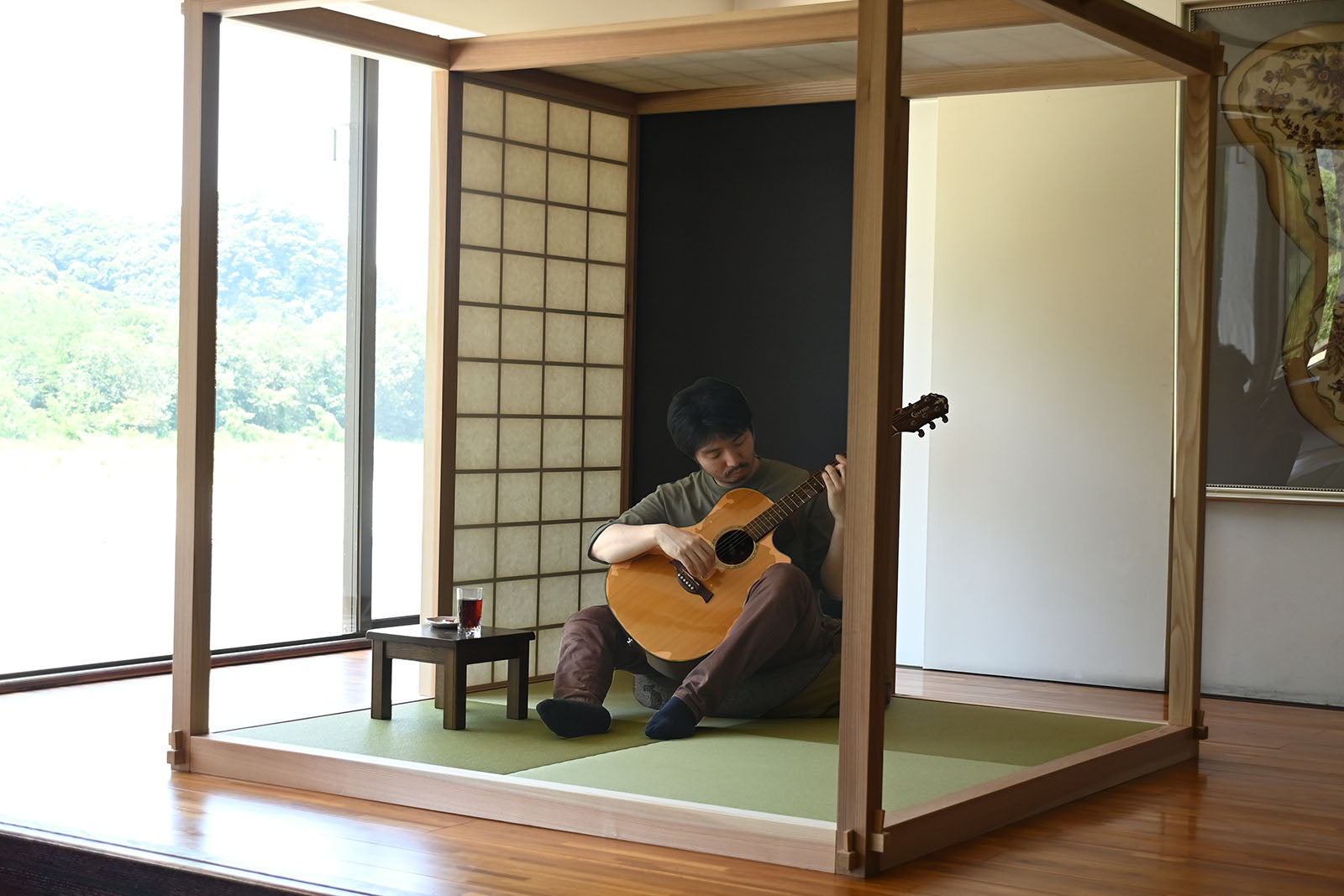 木のフレームで囲まれたRoom内の畳に座り、アコースティックギターを弾く男性。